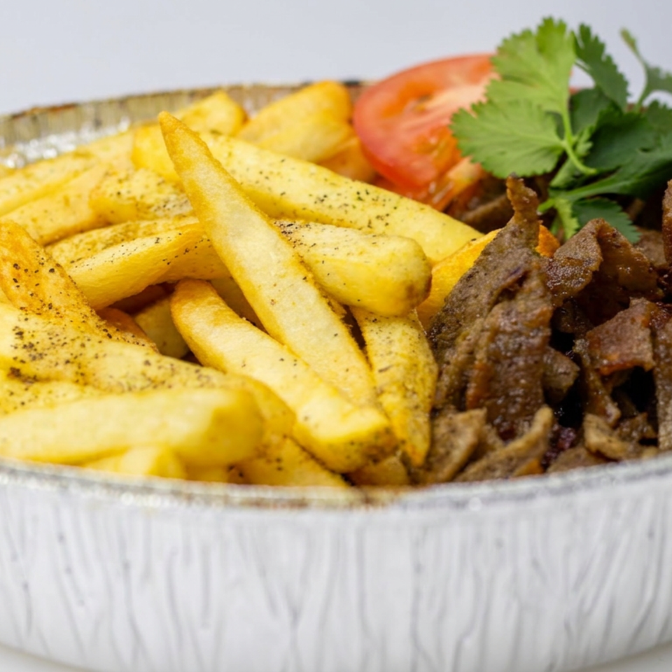 Seasoned French fries and sliced grilled meat served in a foil tray with tomato and fresh parsley garnish.